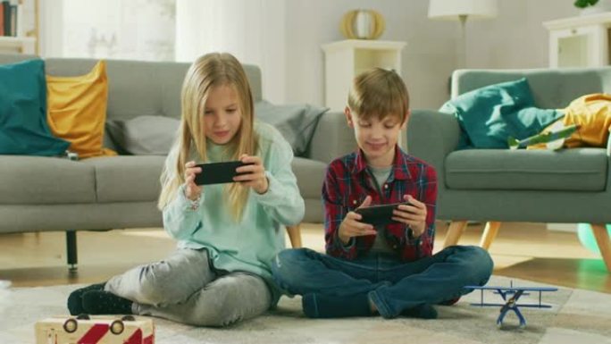 在家: 可爱的小女孩和可爱的男孩在两部智能手机上玩竞争性视频游戏。男孩和女孩每个人都在水平景观模式下