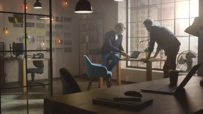 两位年轻英俊的设计师坐在办公桌上，在他们的笔记本电脑上讨论社交媒体应用程序界面。他们在阳光明媚的创意