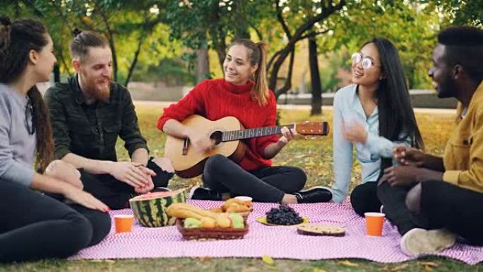 多种族的朋友在公园唱歌，拍手，而美丽的女孩在野餐时弹吉他。食物和毯子是可见的。