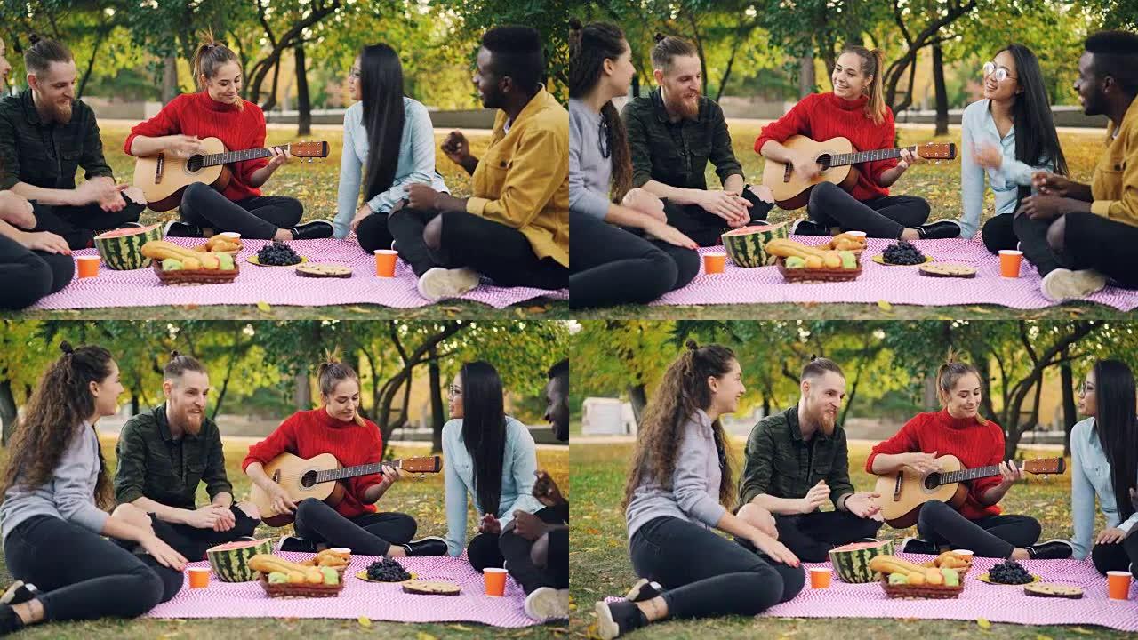多种族的朋友在公园唱歌，拍手，而美丽的女孩在野餐时弹吉他。食物和毯子是可见的。