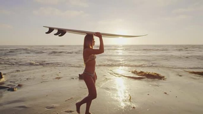 日落时分，冲浪板在头顶上方的沙滩上行走的冲浪女孩