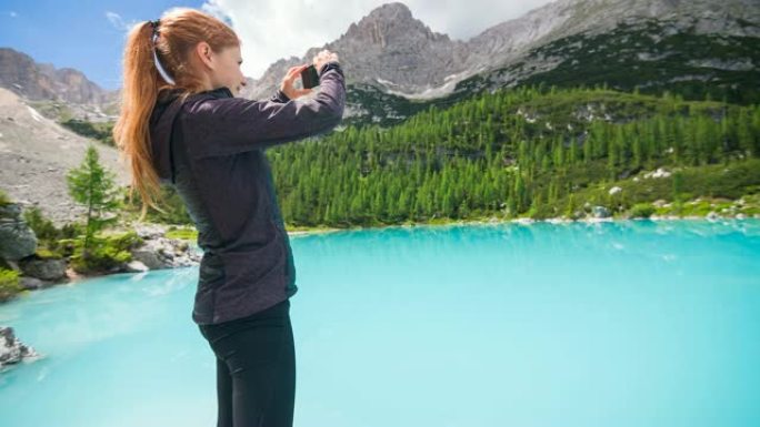 山湖的女游客欣赏美景，用智能手机拍照，背景为山脉