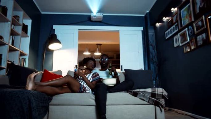 快乐的年轻休闲非洲夫妇坐在家里的沙发上，用饮料和爆米花慢动作在投影仪上看电影。
