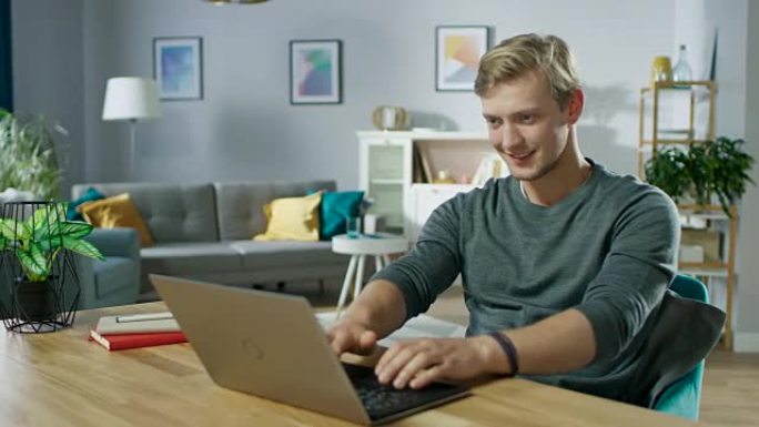 英俊的微笑的年轻人坐在舒适的客厅的桌子旁使用笔记本电脑。