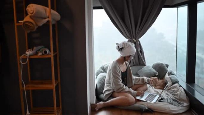 一名亚洲华裔少女在卧室里用笔记本电脑上网，同时用猫做面部水疗面罩