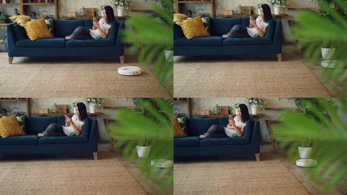 当机器人胡佛在家里的地板和地毯上徘徊时，亚洲学生正在使用坐在沙发上的智能手机。电器、家用和科技概念。