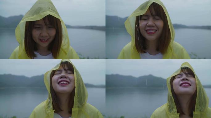 慢动作 -- 年轻的亚洲女人穿着雨衣站在湖边玩雨感到快乐。生活方式女性在雨天享受和放松。