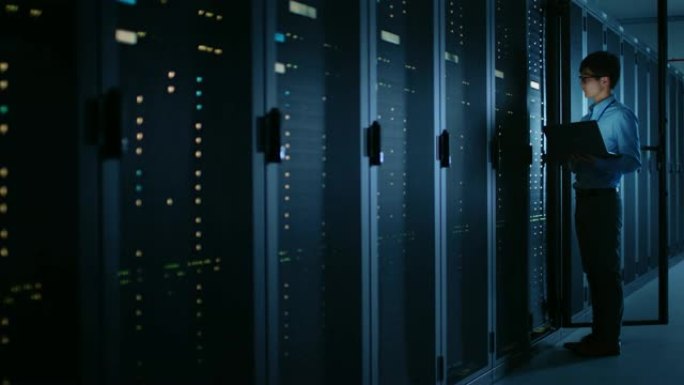 在黑暗的数据中心: 男性IT专家站在一排操作服务器机架旁边，使用笔记本电脑进行维护。云计算、人工智能