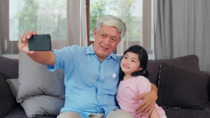 亚洲祖父和孙女在家视频通话。资深的中国爷爷很高兴与年轻女孩使用手机视频通话与躺在客厅里的爸爸和妈妈交