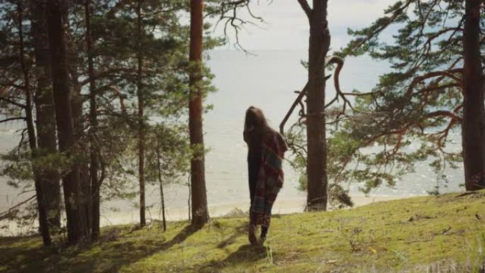 穿着黑色衣服的年轻女子在欧洲松树林中散步。这是大自然温暖的晴天。她来到边缘，看着海景。