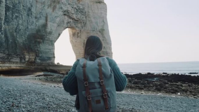 后视快乐的旅游女人，背着背包，沿着美丽的海岸探索白色拱形岩石，靠近埃特雷塔慢动作