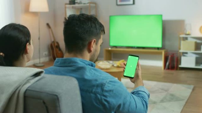 幸福的夫妇坐在家里的客厅里看绿色色度键屏幕电视，在沙发上放松。Guy还使用绿色模拟屏幕智能手机。