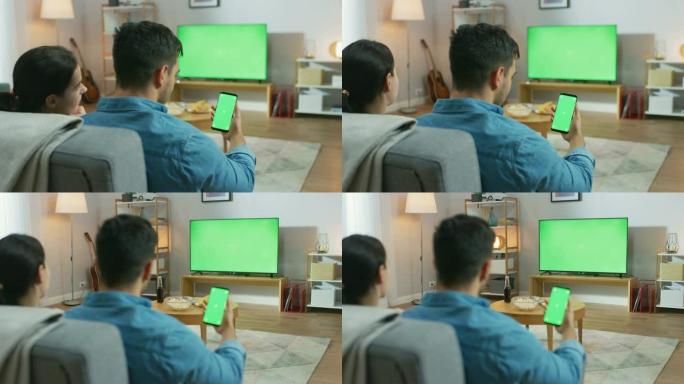 幸福的夫妇坐在家里的客厅里看绿色色度键屏幕电视，在沙发上放松。Guy还使用绿色模拟屏幕智能手机。