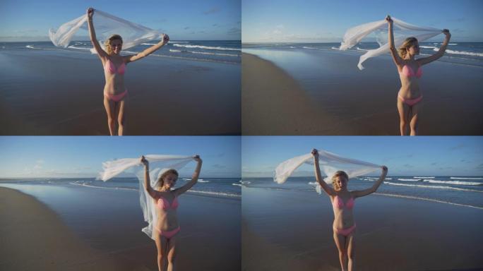南非，一个微笑的美丽年轻女子站在海滩浅水中，拿着白色纱笼，4k慢动作视图