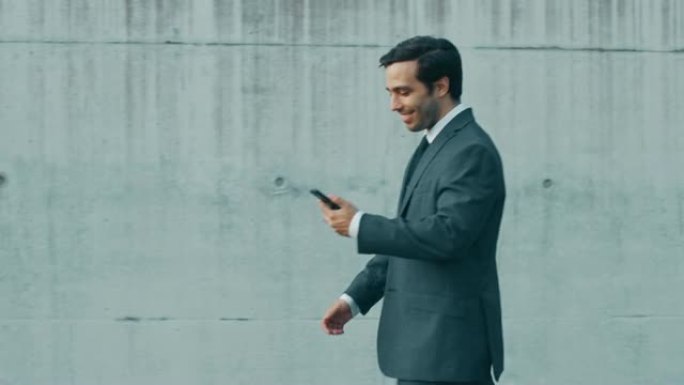 快乐的商人正带着智能手机在城市混凝土墙旁边的街道上行走。他穿着灰色西装。晴天。