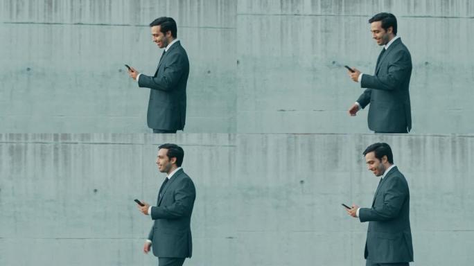 快乐的商人正带着智能手机在城市混凝土墙旁边的街道上行走。他穿着灰色西装。晴天。