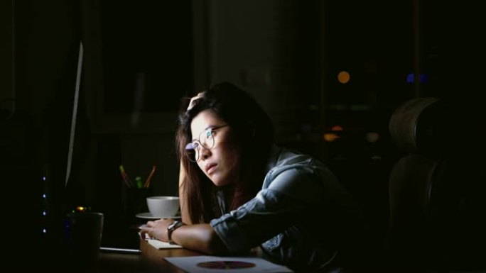 4k镜头的场景有吸引力的亚洲女性工作到很晚，在黑暗中工作场所的计算机显示器桌面前的桌子上认真思考，工