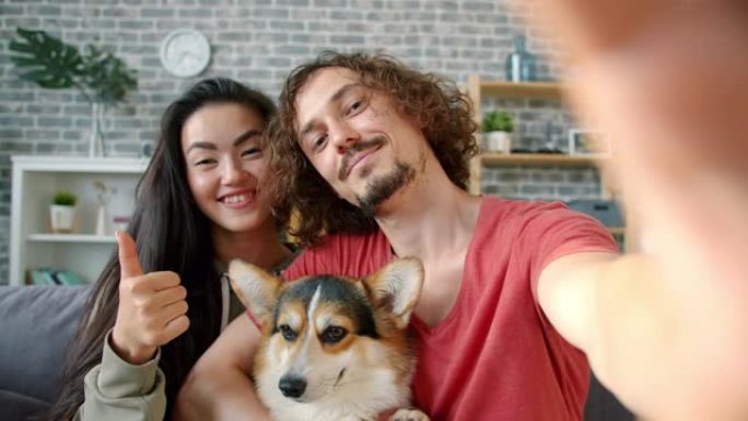 幸福的夫妇在家里与柯基犬自拍的肖像显示竖起大拇指