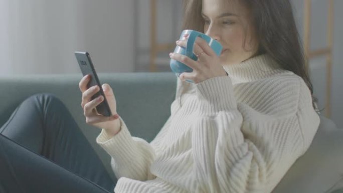 美丽的年轻女子使用智能手机，坐在椅子上喝茶。性感的女孩穿着毛衣上网，在社交媒体上发帖，在舒适的公寓里