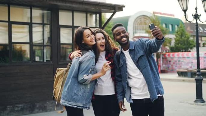 快乐的非洲裔美国人正在与朋友自拍，美丽的高加索女孩站在街上摆姿势，在国外旅行时拿着智能手机。