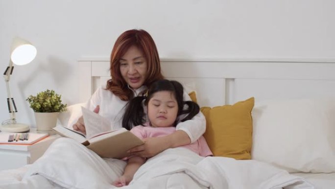 亚洲祖母在家给孙女读童话。高年级的中国人，奶奶很高兴与年轻的女孩一起放松，她晚上睡在家里卧室的床上听
