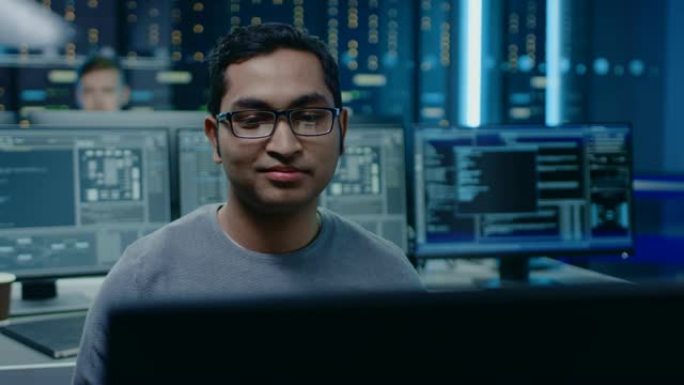 一个聪明英俊的软件开发人员戴着眼镜在个人计算机上工作的肖像，对着镜头微笑。在后台，技术室没有屏幕的个