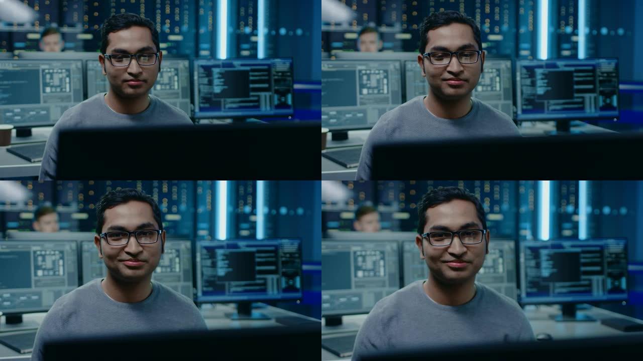一个聪明英俊的软件开发人员戴着眼镜在个人计算机上工作的肖像，对着镜头微笑。在后台，技术室没有屏幕的个