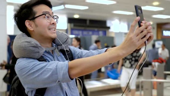 年轻的亚洲背包客博主男子在机场值机柜台自拍，配有旅行包和颈枕