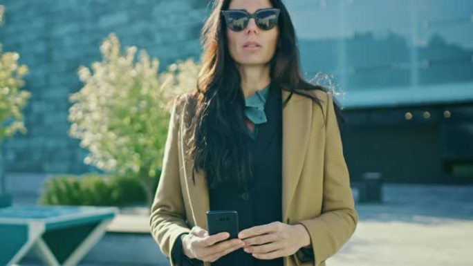 优雅的女商人在现代玻璃建筑前行走时使用智能手机开展业务。穿着外套和墨镜的美丽时尚女人在现代城市环境中