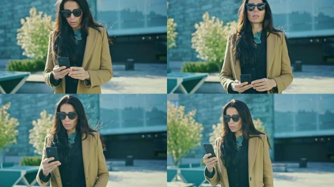 优雅的女商人在现代玻璃建筑前行走时使用智能手机开展业务。穿着外套和墨镜的美丽时尚女人在现代城市环境中