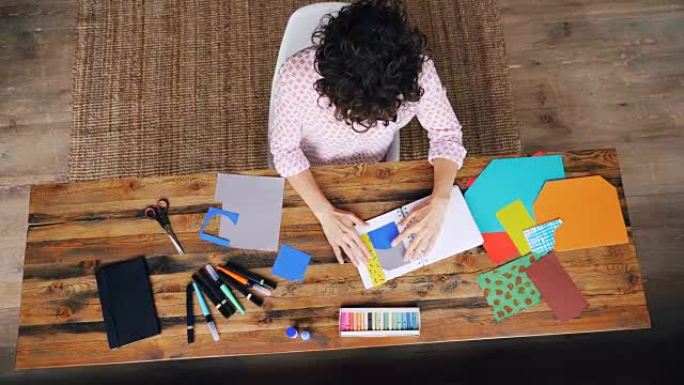 专业设计师在桌子上制作拼贴画的俯视图，用剪刀将图形粘贴在笔记本上并切割形状。工作场所和创造力概念。