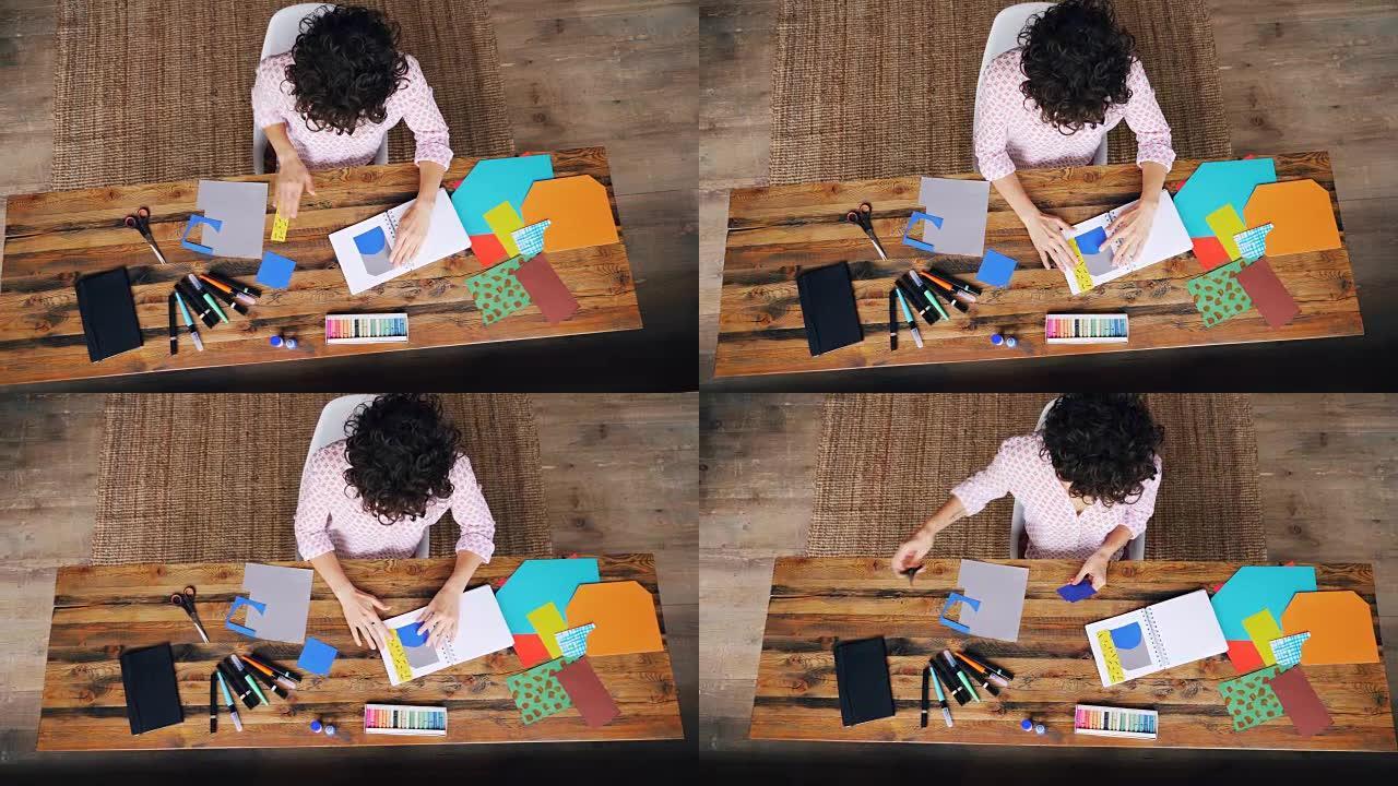专业设计师在桌子上制作拼贴画的俯视图，用剪刀将图形粘贴在笔记本上并切割形状。工作场所和创造力概念。