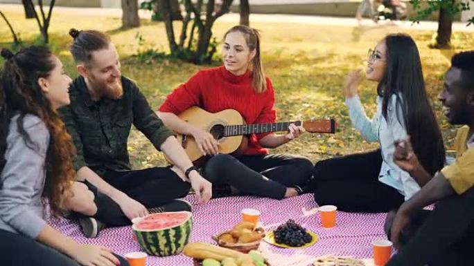 漂亮的年轻女士正在弹吉他，而她的朋友们正在公园里唱歌和听音乐。毯子上可以看到食物水果和糕点。
