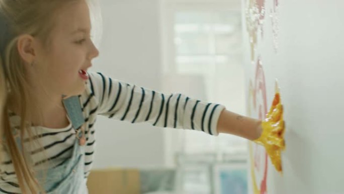 快乐的小女孩用黄色和红色油漆蘸手，开始在墙上画手印。她玩得很开心，笑得很开心。家正在装修。