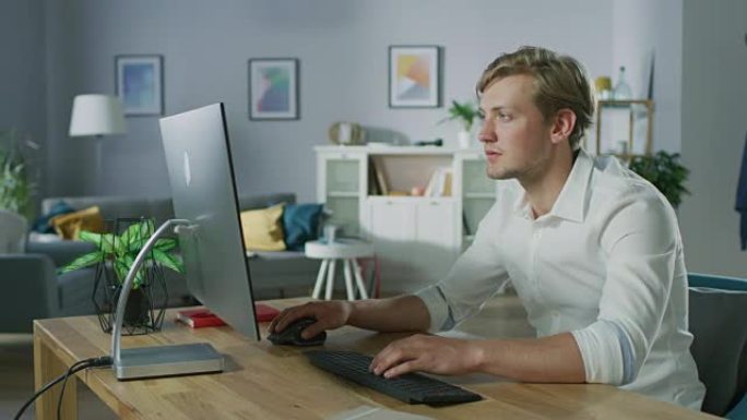 英俊的年轻设计师坐在办公桌前的个人电脑上工作。背景时尚家居装饰。