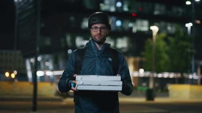 在市中心的一个晚上，一个年轻的披萨快递员在镜头前对自己的工作满意地微笑着