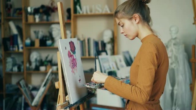 迷人的苗条女孩正在用画笔和调色板的油画画花，并独自在工作室工作。创造力和艺术概念。
