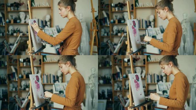 迷人的苗条女孩正在用画笔和调色板的油画画花，并独自在工作室工作。创造力和艺术概念。