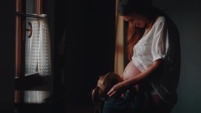 一个可爱的小女儿的真实镜头是亲吻母亲怀孕的肚子，她在母亲的肚子上感到婴儿，并等待她的弟弟或妹妹的出生