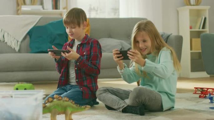 在家中坐在地毯上: 可爱的小女孩和可爱的男孩在两部智能手机上的视频游戏中一起玩，将它们保持在水平景观