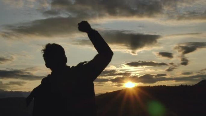一个成功的快乐旅行男人站在欢乐之上，举起手来。快乐的剪影30年代的男人看着山脉后面的日落天空。冒险旅