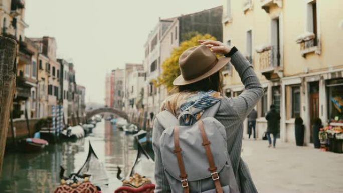 戴着帽子的快乐年轻时尚欧洲女性游客喜欢站在意大利著名的威尼斯运河旁四处张望。
