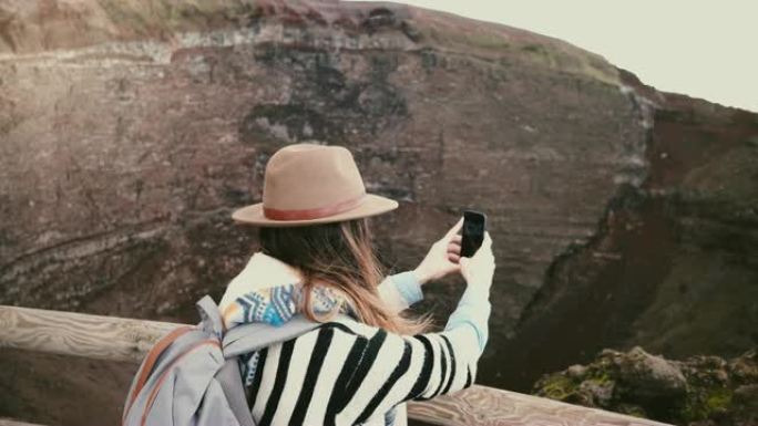 年轻快乐的自由职业者女孩带着背包在意大利度假时拍摄维苏威火山火山口的智能手机照片。