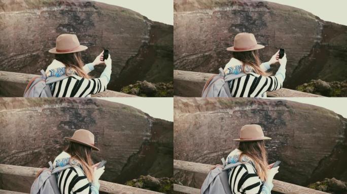 年轻快乐的自由职业者女孩带着背包在意大利度假时拍摄维苏威火山火山口的智能手机照片。