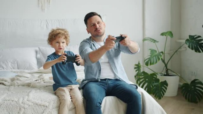 男人和男孩玩电子游戏的慢动作坐在家里的床上玩得开心