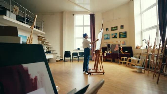 苗条的女人正站在明亮宽敞的工作室中心的画布上绘画，独自创作杰作。创造性工作、职业和职业概念。