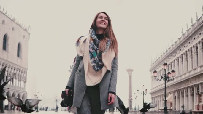快乐美丽的女游客带着相机走在威尼斯慢动作的老城广场上，周围是一群鸽子。