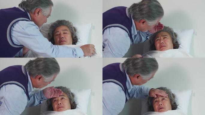 年迈的丈夫正在家里的床上照顾生病的妻子。通过用手触摸妻子的额头来测量发热，并注意不远处的症状