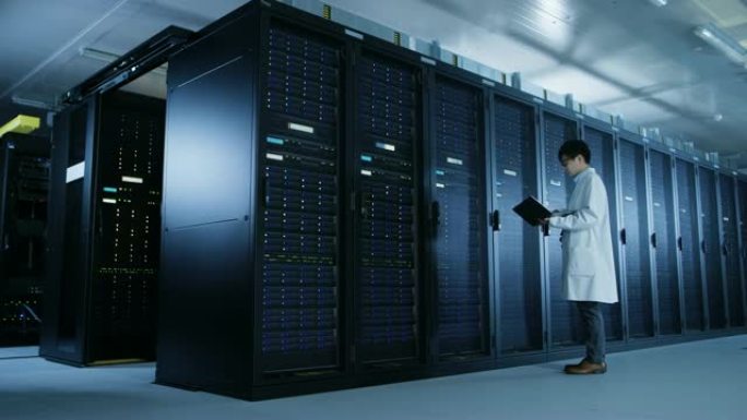 在数据中心: 穿着白大褂的男性IT专家走在一排服务器机架旁边，使用笔记本电脑运行维护诊断工具，进行控