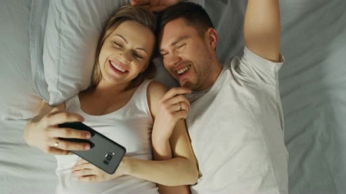 充满爱心的年轻夫妇在床上度过了早晨，怀孕的年轻女子在触摸屏智能手机上向伴侣展示了一些东西，并在社交网